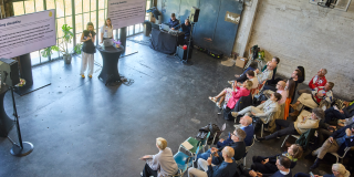 &Aacute;ine Aherne stands in front of a large seated crowd with a microphone. There are two screens on either side of her that read "Defining disability"